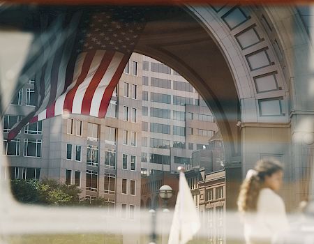 A large American flag hangs inside a grand arched courtyard, framed by white stone buildings and city rooftops beyond.