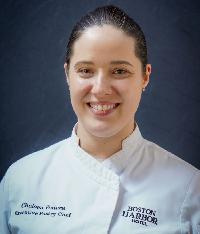 A female executive pastry chef in a white jacket with name/titles, smiling for a headshot against a blue backdrop.
