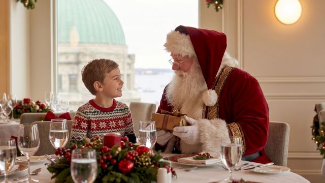 A child in a cozy sweater chats with Santa in a festive dining room, with Christmas lights and gifts on the table.