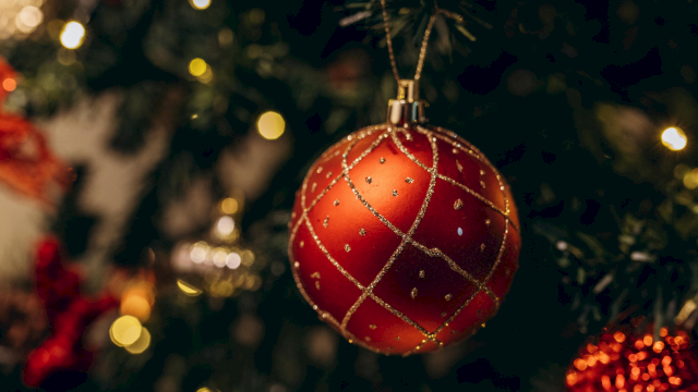 A red jeweled Christmas bauble hangs on a decorated tree, with warm holiday lights softly blurred in the background.