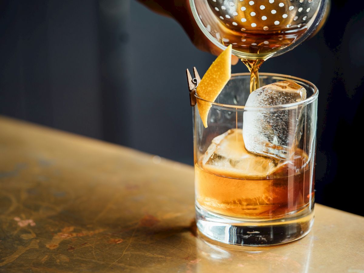Amber liquor is being poured into a rocks glass over ice, with a citrus slice balanced on the rim.