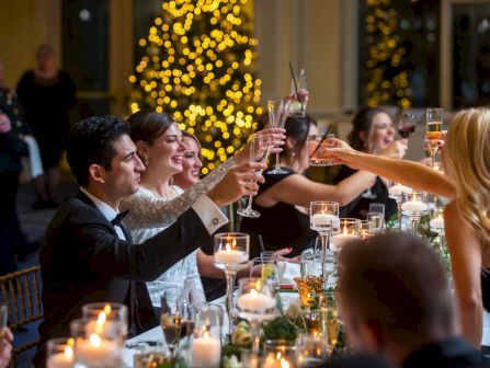 Guests in formalwear raise champagne flutes at a candlelit dinner table, with sparkling holiday lights behind them.