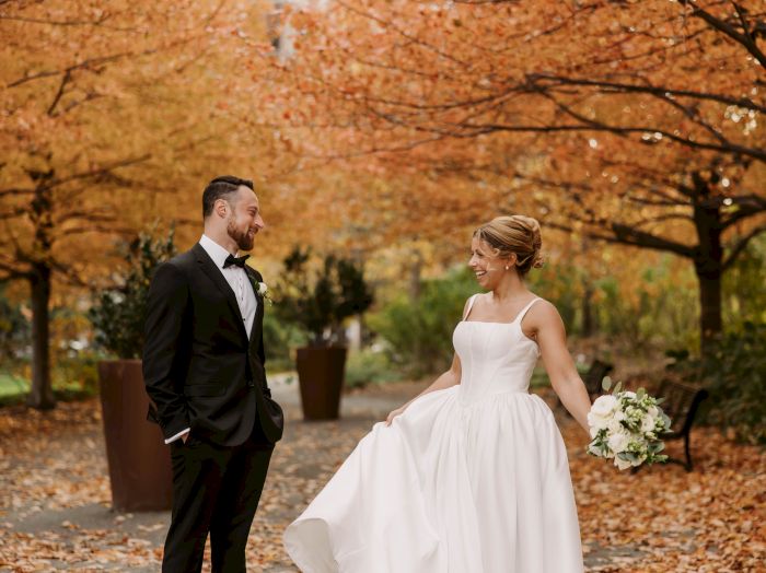 A bride in a white gown and a groom in a tux share a joyful moment outdoors amid autumn leaves, holding flowers.