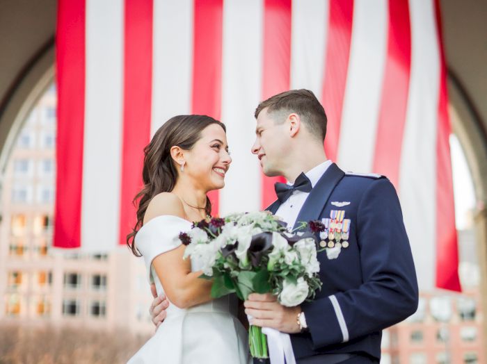 A bride and groom smile in front of a huge American flag backdrop during a wedding ceremony, holding a bouquet.