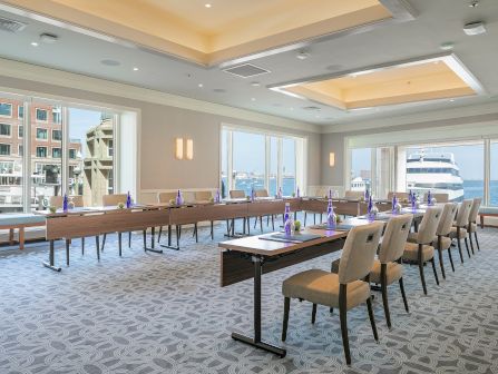 A modern conference room set with long tables, chairs, and blue bottles, with windows overlooking a harbor and a large ship.