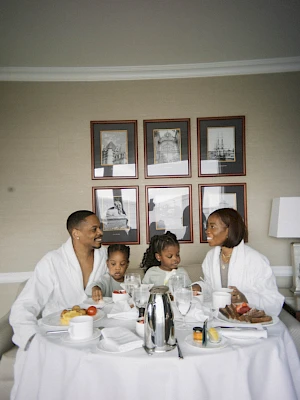 A family enjoys breakfast in a hotel room, wearing robes and seated around a table with framed pictures on the wall behind them.