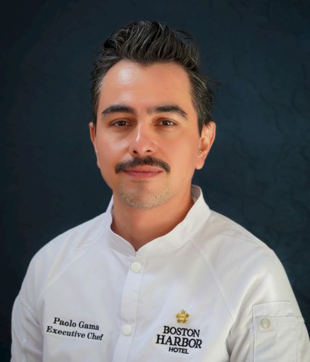 A male executive chef portrait in a white jacket labeled Paolo Gama, Boston Harbor Hotel, against a dark backdrop.