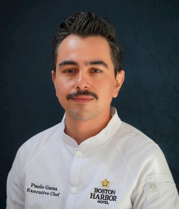 A male executive chef portrait in a white jacket labeled Paolo Gama, Boston Harbor Hotel, against a dark backdrop.