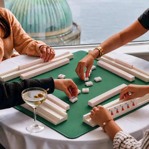 People play a tabletop domino-style game on green felt near a glass of wine, with several hands arranging tiles around the table.