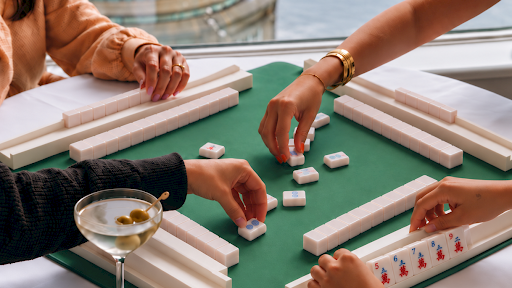 People play a tabletop domino-style game on green felt near a glass of wine, with several hands arranging tiles around the table.