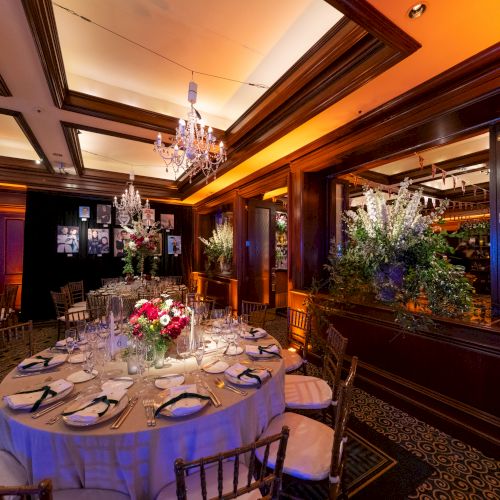 An elegant dining room with round tables set for an event, featuring chandeliers, floral arrangements, and wooden decor.