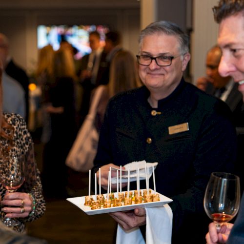 People at an event are socializing. A server is offering appetizers on a tray while holding glasses, and everyone seems engaged.