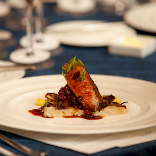 A gourmet dish features a piece of meat on a bed of creamy risotto, garnished with herbs and vegetables on a white plate.
