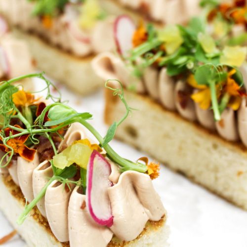 Garnished finger sandwiches with whipped filling, radish slices, greens, and edible flowers are displayed on a white surface.
