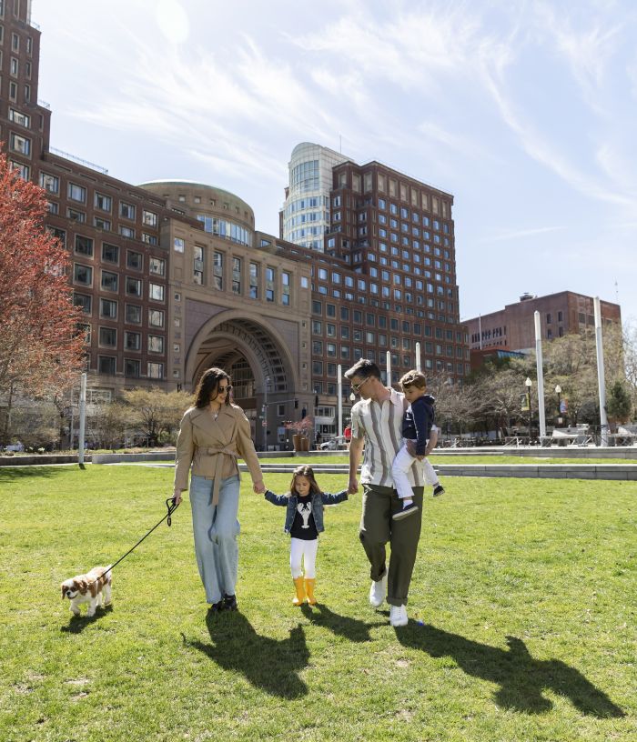 A family walks hand-in-hand with a toddler and a leashed dog on a lawn outside tall brick buildings under a blue sky.