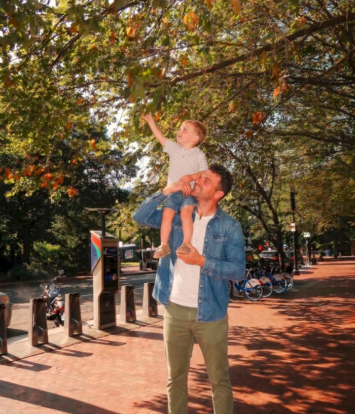 A dad lifts his little child in the air under leafy trees in a sunny street, surrounded by scooters and passing cars.