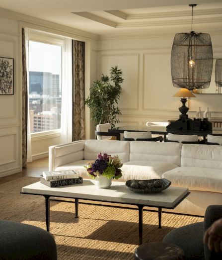 A bright living room with a white sofa, marble coffee table, flower bouquet, hanging pendant lights, and a leafy plant by the window.