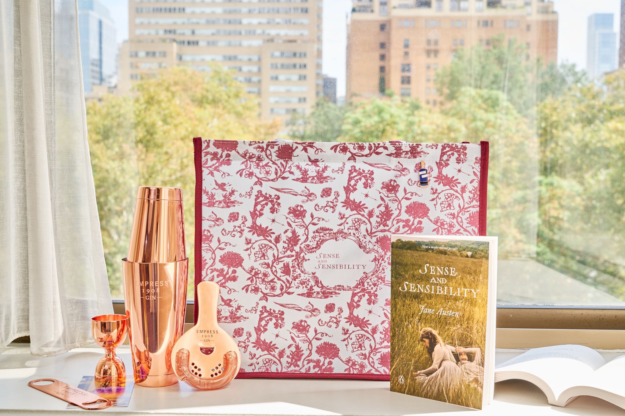 A desk by a window shows copper gin items, a glass, and a red patterned bag beside the &ldquo;Sense and Sensibility&rdquo; book.