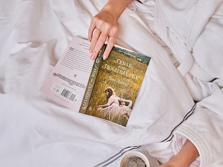 A woman relaxing on white bedding in a robe reads *Sense and Sensibility* while holding a coffee by a croissant.