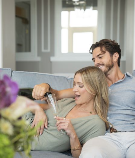 A couple lounges on a couch, smiling as they pour champagne into glasses for a toast in a bright living room.