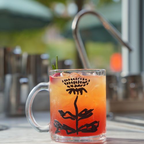 A clear mug with a colorful orange-red iced drink, garnished with a straw, set on a patio with soft bokeh lights.