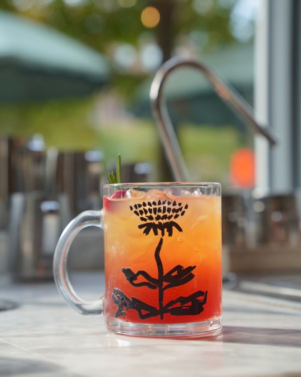 A clear mug with a colorful orange-red iced drink, garnished with a straw, set on a patio with soft bokeh lights.