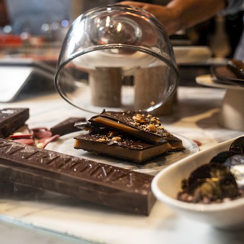 A chef pours melted chocolate from a metal bowl onto a table covered with chocolate pieces and chopped nuts in a caf&eacute; kitchen.