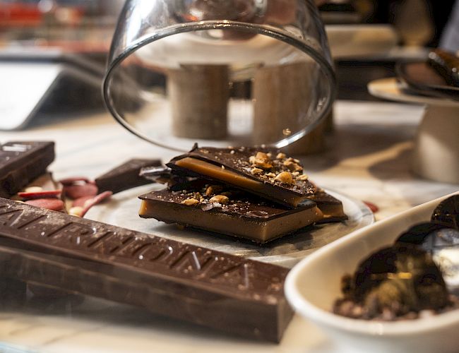 A chef pours melted chocolate from a metal bowl onto a table covered with chocolate pieces and chopped nuts in a caf&eacute; kitchen.
