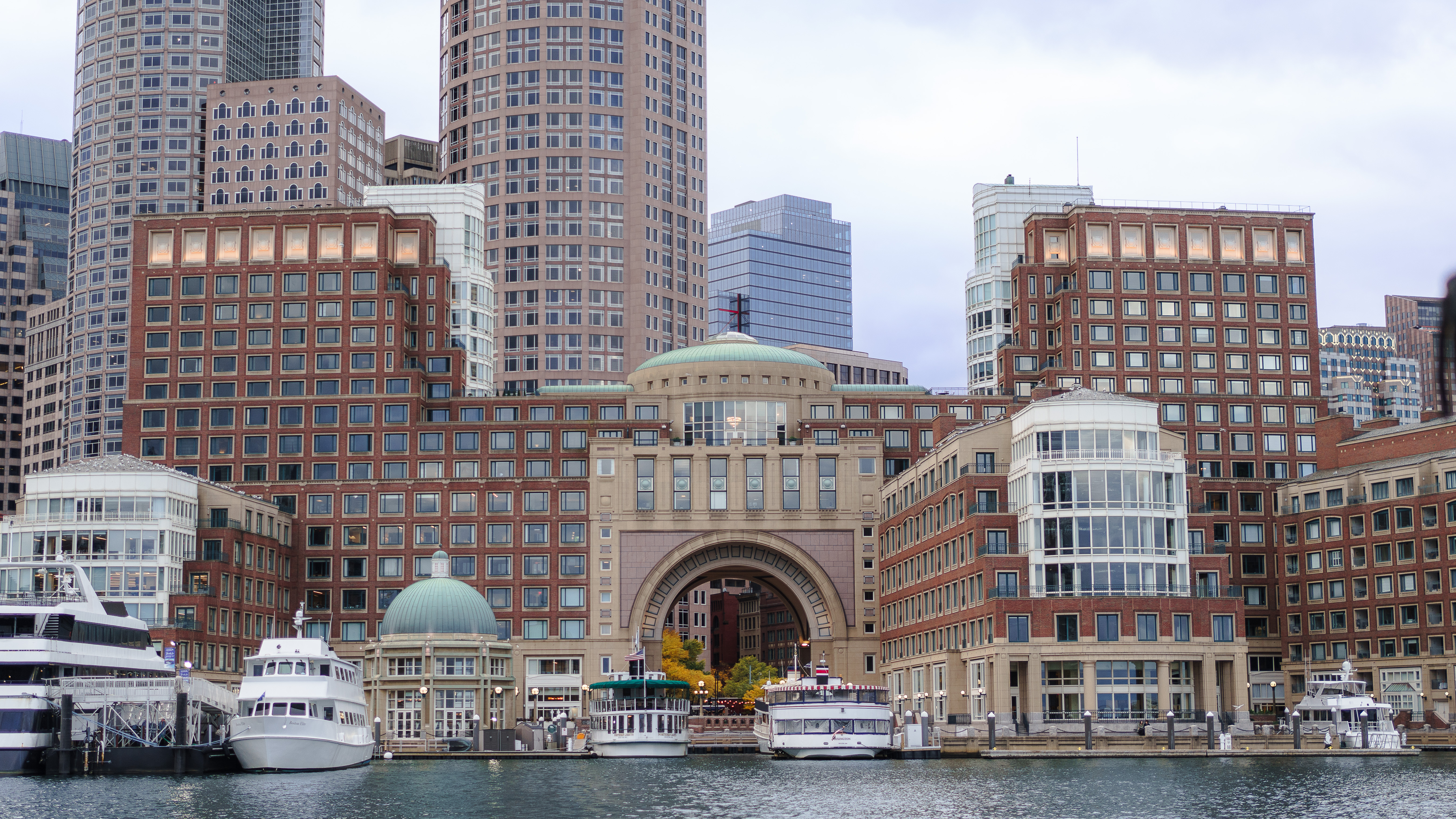 A busy harbor with docked boats sits in front of red-brick and glass office buildings in a downtown cityscape.