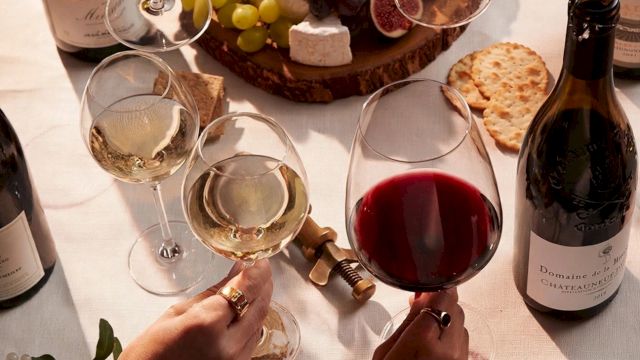 Friends clink glasses of red and white wine over a cheese board of grapes, figs, and crackers during a toast.