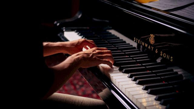 A person&rsquo;s hands play a piano, pressing the keys in a dimly lit studio with sheet music in view.