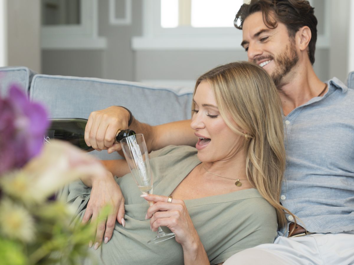 A smiling couple relaxes on a couch, with one person pouring a drink into a champagne flute for the other.