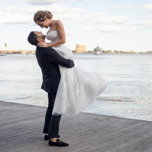A couple is by the water; the person in a suit lifts the one in a wedding dress on a pier, with boats and skyline in the background.