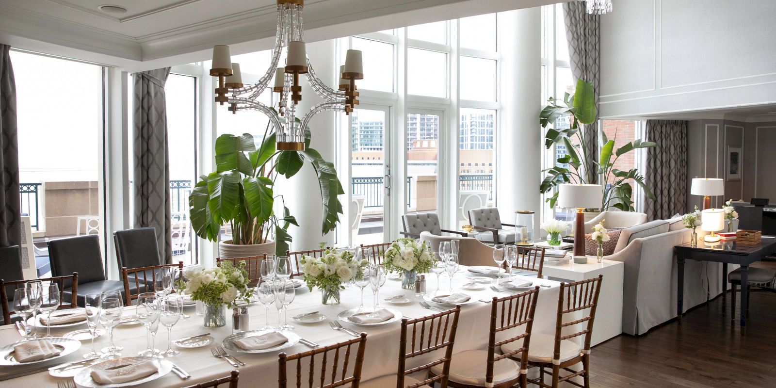 Elegant dining room with a long table set for a formal meal, featuring plants, chandeliers, and large windows with city views.