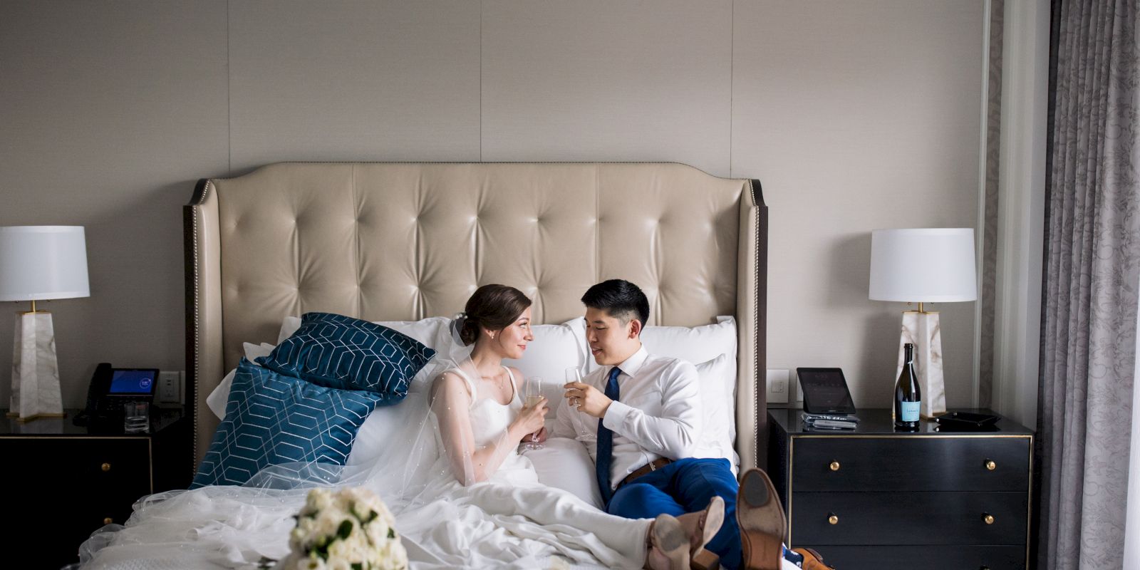 A couple is sitting on a bed in a well-decorated room, holding hands, with bedside tables featuring lamps on either side of the bed.