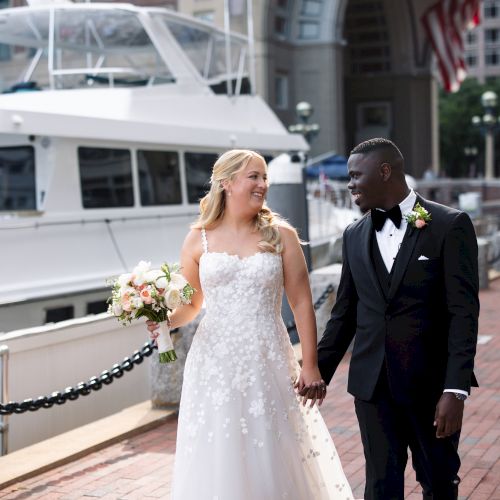 A couple in wedding attire is walking hand-in-hand by a waterfront, with a yacht in the background.