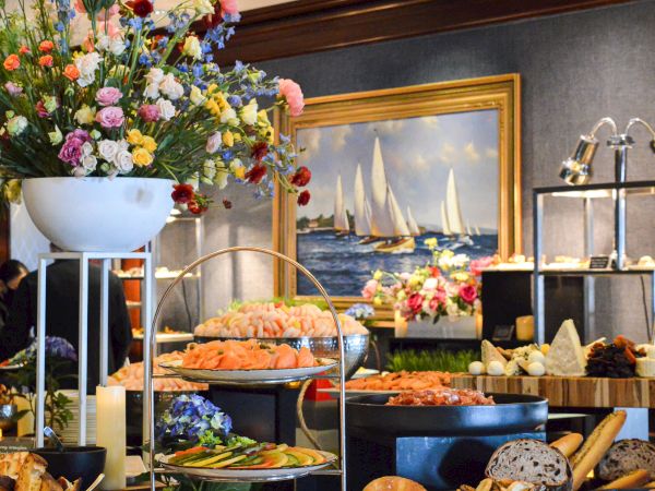A buffet setup with assorted foods such as seafood, cheese, and bread is adorned with floral arrangements and a sailboat painting in the background.