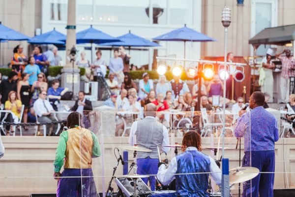 A band performing outdoors with an audience seated under blue umbrellas. Bright stage lights illuminate the scene.