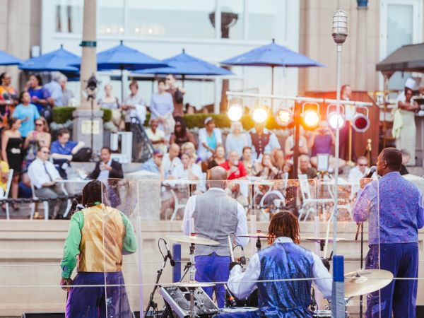 A band performing outdoors with an audience seated under blue umbrellas. Bright stage lights illuminate the scene.