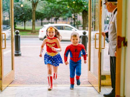 Two kids in superhero costumes (Wonder Woman and Spider-Man) run happily through open doors, a smiling hotel lobby attendant watches.