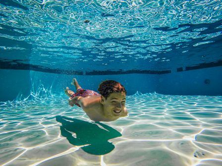 A person swimming underwater in a pool, with sunlight creating patterns on the pool floor and a joyful expression on their face.