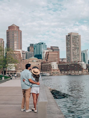 A couple stands on a waterfront boardwalk, embracing while looking at a city skyline with tall buildings under a partly cloudy sky.