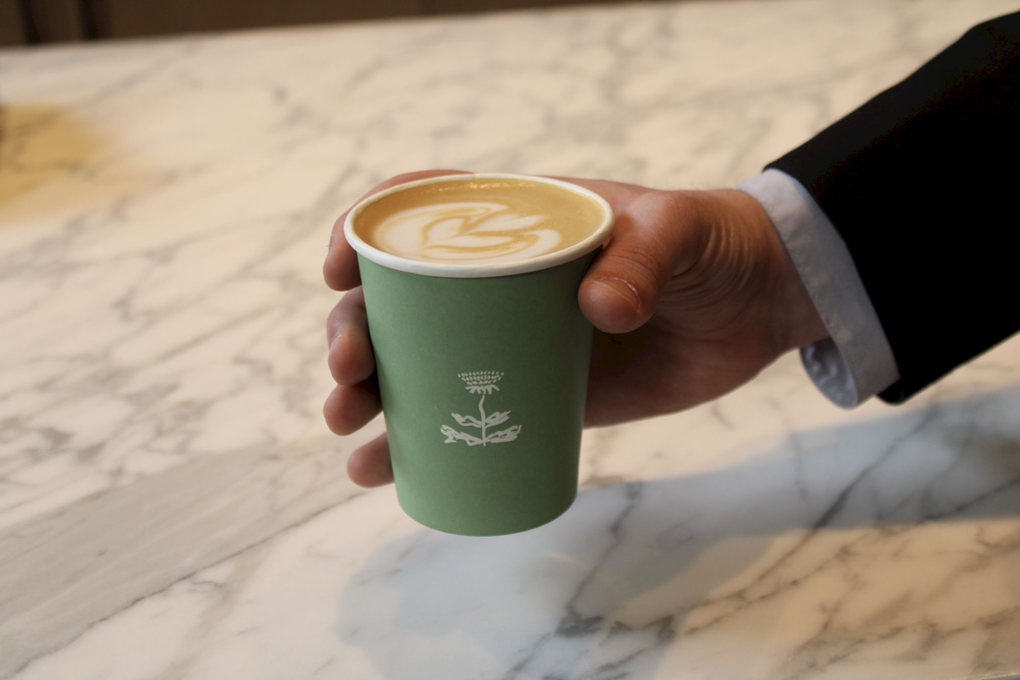 A hand holding a green paper cup of coffee with latte art on a marble table.