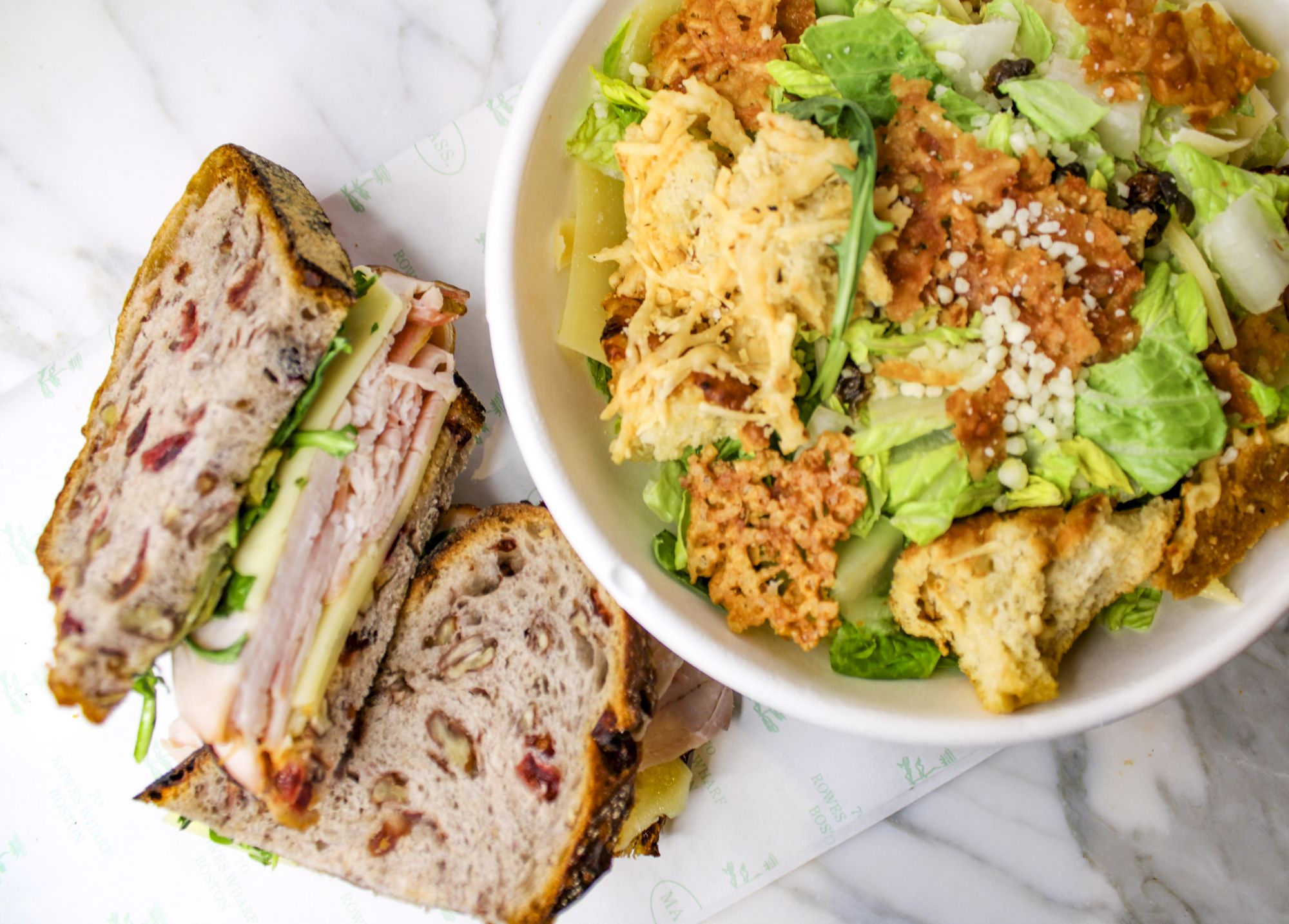 A sandwich with deli meat, cheese, and greens on multigrain bread is next to a salad with lettuce, chicken, dressing, and croutons.