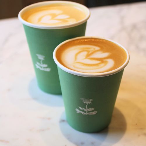 Two cups of coffee with latte art in green paper cups are placed on a marble table.