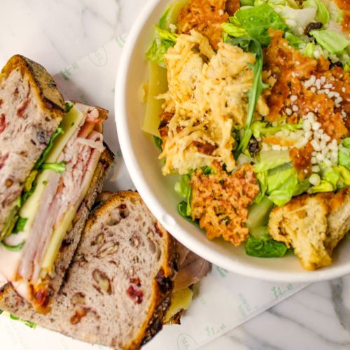 A sandwich with cold cuts and cheese on seeded bread, next to a salad with lettuce, chicken, and dressing in a bowl.