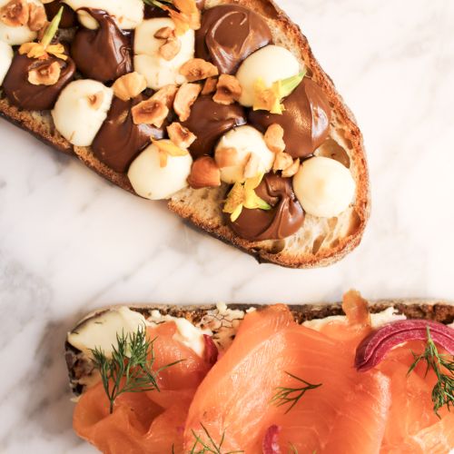 Two pieces of toast: one with chocolate, nuts, and fruit; the other with smoked salmon, red onion, and dill on a marble surface.