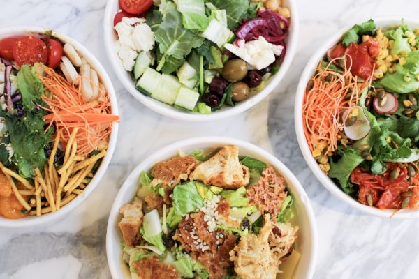 Four bowls of salads with various ingredients like greens, olives, cherry tomatoes, cucumbers, shredded carrots, and croutons are shown.