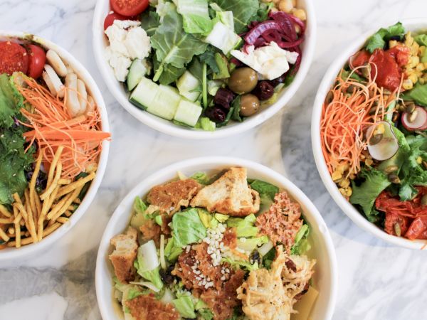 Four bowls of salads with various ingredients like greens, olives, cherry tomatoes, cucumbers, shredded carrots, and croutons are shown.