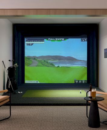 The image shows a golf simulator setup with a screen displaying a golf course, two chairs, a side table, and golf equipment in a cozy room.